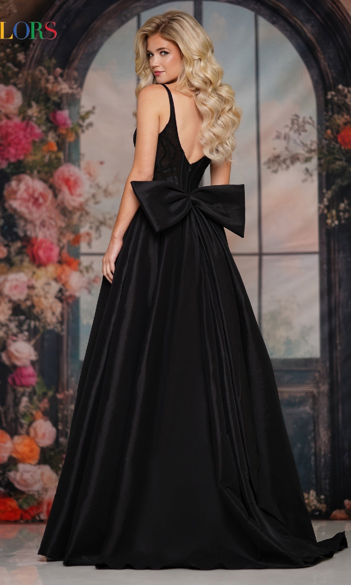 A woman with long, wavy blonde hair models the Colors Dress Statement-Bow Long Prom Dress (Style 3922), a sleeveless black gown with a deep V-back and large waist bow, posing before floral décor and an arched window.